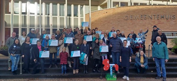 Op de foto staat wethouder Rieja Raven samen met 45 winnaars van MOEK 2024 op het bordes van het gemeentehuis. De winnaars hebben in hun hand een checque waarop het bedrag staat dat ze hebben gewonnen.